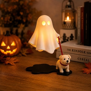Op de afbeelding: Een witte, spookvormige lamp met twee ronde oogopeningen, van binnenuit verlicht. Het spook is verbonden via een rode riem aan een klein, crèmekleurig puppybeeldje met een rode halsband, beide gemonteerd op een zwarte voet.