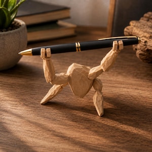 May include: A light brown, carved wooden figurine with a faceted texture, shaped like a figure holding a black ballpoint pen with gold-coloured accents horizontally above its head. The pen features a gold tip, a gold clip, and gold rings.
