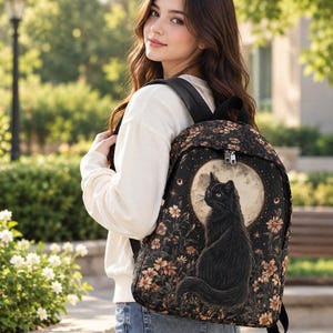 Puede incluir: Una mochila negra con un estampado gráfico de un gato negro sentado frente a una luna grande y pálida, rodeado de pequeñas flores naranjas y estrellas. La mochila tiene correas negras, un compartimento principal con cremallera y forma estructurada.