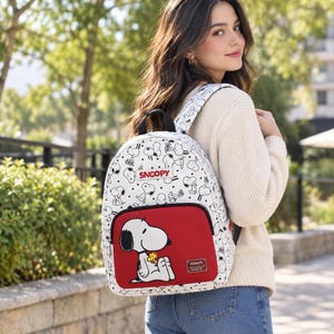 Könnte beinhalten: Ein weißer Rucksack mit einem durchgehenden schwarz-weißen Druck von Snoopy-Figuren. Die Vordertasche ist einfarbig rot und zeigt eine große Grafik von Snoopy, der Woodstock hält. Ein rechteckiger Aufnäher oben zeigt SNOOPY und ein Metallschild PEANUTS.
