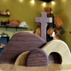 May include: A wooden cross stands tall above three wooden hills, two of which are brown and one is a light beige. The hills have rounded tops and openings, resembling a tomb.