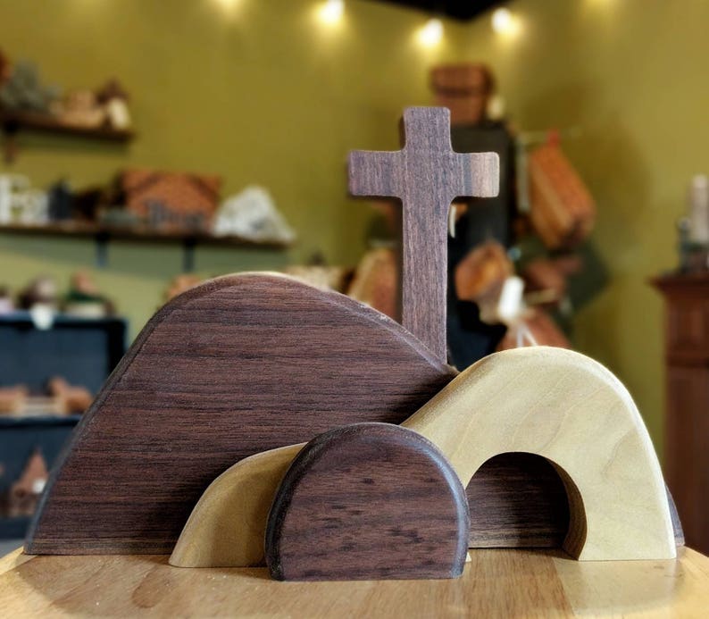 May include: A wooden cross stands tall above three wooden hills, two of which are brown and one is a light beige. The hills have rounded tops and openings, resembling a tomb.