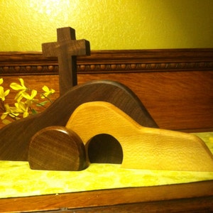 May include: A wooden Easter tomb set with a cross, a brown hill, and a light brown tomb with an opening. The set is on a green surface with yellow flowers.