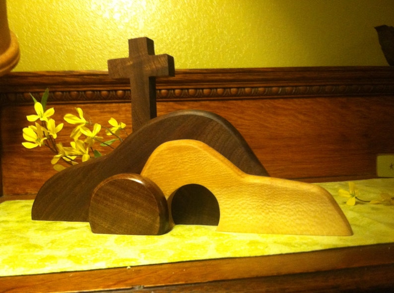 May include: A wooden Easter tomb set with a cross, a brown hill, and a light brown tomb with an opening. The set is on a green surface with yellow flowers.