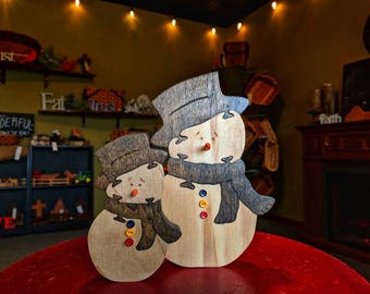 May include: Two wooden snowman ornaments with painted details. The larger snowman is wearing a brown scarf and has a black hat. The smaller snowman is wearing a brown scarf and has a black hat. Both snowmen have black eyes and red noses.