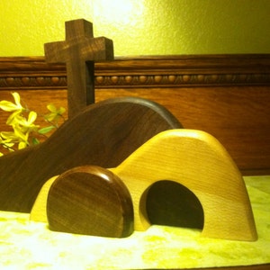 May include: A wooden Easter tomb with a cross on top. The tomb is made of two pieces of wood, one brown and one light brown. The tomb is open, and the cross is standing upright. There are yellow flowers in the background.
