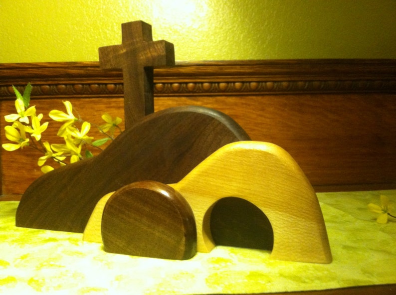 May include: A wooden Easter tomb with a cross on top. The tomb is made of two pieces of wood, one brown and one light brown. The tomb is open, and the cross is standing upright. There are yellow flowers in the background.