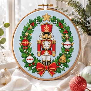 Può includere: Un telaio da ricamo circolare in legno contenente un motivo a punto croce di uno schiaccianoci centrato all'interno di una ghirlanda di agrifoglio verde. La ghirlanda è decorata con ornamenti rossi, palline bianche con cuori rossi e una campana.