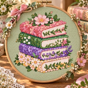 May include: A circular wooden embroidery hoop containing a cross-stitch pattern of a stack of four books on a light green fabric. The books are decorated with pink, green, purple, and white flowers. The hoop is adorned with pink ribbon and flowers.