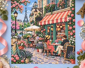 Può includere: Un ricamo a punto croce che raffigura una scena di strada parigina con un caffè, una tenda a righe, fiori rosa, una bicicletta e la Torre Eiffel in lontananza. L'opera mostra due persone sedute a un tavolo, un lampione e un sentiero acciottolato, realizzati con fili dai colori vivaci e dettagliati.