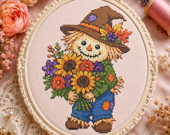 Può includere: Un ricamo a punto croce in una cornice ovale raffigurante uno spaventapasseri con un cappello marrone decorato con foglie autunnali, una camicia verde e una salopette blu. Lo spaventapasseri tiene in mano un grande bouquet di girasoli e fiori.