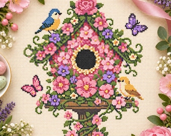 Op de afbeelding: Een kruissteekpatroon op crèmekleurige stof met een vogelhuisje bedekt met roze en paarse bloemen. Een blauwe vogel zit op het dak, een gele vogel rust op de zitstok en twee paarse vlinders bevinden zich aan de zijkanten van de structuur.