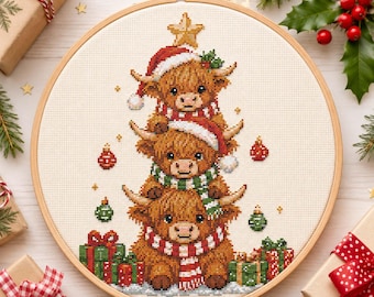 May include: A cross-stitch embroidery in a round wooden hoop featuring three stacked Highland cows wearing red and white Santa hats and striped scarves. The design includes a yellow star on top, red and green ornaments, and wrapped gift boxes.