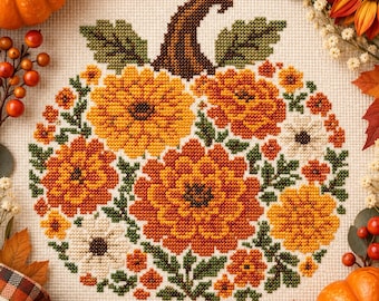 Può includere: Un ricamo a punto croce su tessuto color crema raffigurante una zucca composta da vari fiori gialli, arancioni e bianchi con foglie verdi e un gambo marrone scuro, circondato da zucche vere, pigne e foglie autunnali.