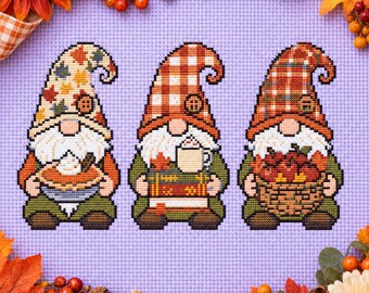 May include: A cross-stitch pattern on light purple fabric featuring three gnomes. The first wears a leaf-print hat and holds a pie. The second wears a plaid hat and holds a mug and books. The third wears a plaid hat and holds a basket of apples.