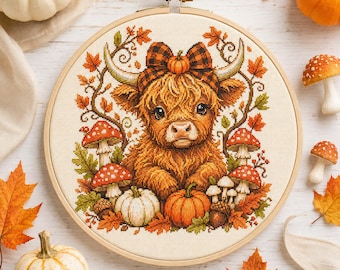 May include: A cross-stitch embroidery in a circular wooden hoop featuring a fluffy Highland cow with a plaid bow, surrounded by autumn leaves, red and white spotted mushrooms, and orange and white pumpkins on a cream-coloured fabric background.