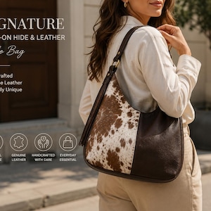 Pode incluir: Uma bolsa hobo que combina pele com pelo em tons de castanho e branco com couro castanho escuro sólido. A bolsa inclui uma alça de ombro com ferragens metálicas e uma borla de couro. O texto na imagem diz SIGNATURE HAIR-ON HIDE & LEATHER.