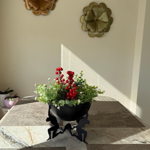 May include: A black bowl planter supported by three stylized human figures in a kneeling position, filled with artificial greenery and bright red berries. The bowl has a smooth matte finish and a rounded shape, creating a decorative centerpiece.