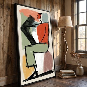 Op de afbeelding: Een ingelijst rechthoekig wandkunstwerk met een gestileerde abstracte figuur in zwarte inktlijnen. De compositie bevat gedurfde kleurvlakken in oranje, saliegroen, gedempt geel en oudroze op een crèmekleurige achtergrond.