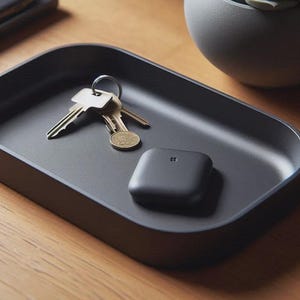 May include: A matte black rectangular valet tray with rounded corners containing a set of silver metal keys on a ring and a small square black electronic tracker device with a centred logo icon, resting on a flat surface.