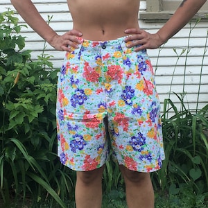 Op de afbeelding: Korte broek met bloemenprint op een lichtblauwe achtergrond, met rode, gele en paarse bloemen. De korte broek wordt gecombineerd met een zwarte crop top en witte sneakers. Een zwarte hoed wordt op het hoofd gedragen.