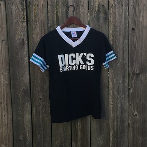 May include: Black athletic t-shirt with white collar and sleeve stripes. The shirt features the text "DICK'S SPORTING GOODS" in white block letters. The shirt is hanging on a black hanger against a wooden fence.