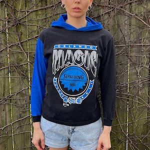 Op de afbeelding: Een persoon draagt een zwart en blauw langarm shirt met de tekst "Orlando Magic" en een Spalding NBA-logo. Het shirt heeft een blauw en zwart kleurblok ontwerp. De persoon draagt spijkerbroek en witte sneakers.