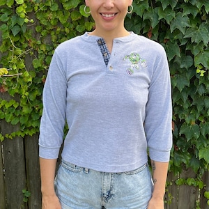Op de afbeelding: Een persoon draagt een lichtgrijs, lang mouwig shirt met een klein geborduurd ontwerp op de voorkant. Het shirt heeft een knoopsluiting en een enkele knoop aan de voorkant. De persoon draagt ook lichtblauwe denim shorts.