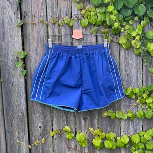 May include: Blue athletic shorts with white and teal trim hanging on a wooden fence covered in green vines. The shorts have an elasticated waistband and are made of a lightweight material. The shorts are displayed outdoors in natural light.