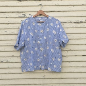 Puede incluir: Camisa de manga corta con estampado de cuadros azules y blancos y estampado floral de margaritas blancas. La camisa está colgada en una percha de madera contra una pared de madera clara y desgastada. Cuello clásico y cierre de botones.