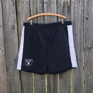 May include: Black athletic shorts with white side panels and a Raiders logo. The shorts are hanging on a wooden hanger against a wooden fence.