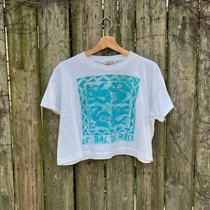 May include: White crop top with a turquoise and blue graphic design. The shirt has the text "LE BALI BALI" printed on it. The shirt is hanging on a wooden fence.
