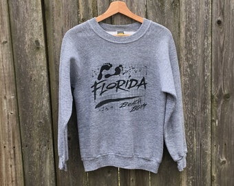 Puede incluir: Una sudadera gris con la palabra "FLORIDA" en negro, junto con un gráfico de huellas y las palabras "Beach Bum". La sudadera está colgada en una valla de madera.