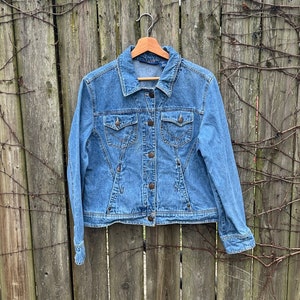 May include: A light blue denim jacket with a classic design, featuring button closures, chest pockets, and a tailored fit. The jacket is hanging on a wooden hanger against a weathered wooden fence.