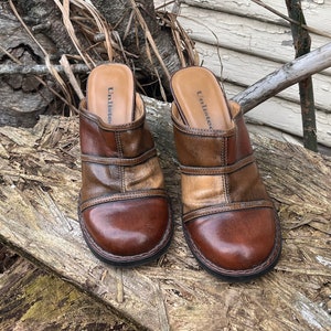 Op de afbeelding: Een paar bruine leren klompen met een blokhak. De klompen hebben een ronde neus en een band over de bovenkant van de voet. Het leer is een mix van lichte en donkere bruintinten. De klompen staan op een houten plank.