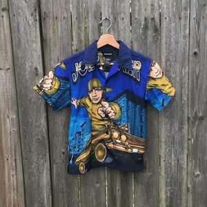 Könnte beinhalten: Kurzärmeliges Button-Down-Hemd mit einem lebendigen Grafikdruck. Das Hemd hat einen blauen Hintergrund mit einer Cartoon-Illustration einer Person und eines Lowrider-Autos. Die Ärmel und der Kragen sind dunkler blau, und der Text "JEEP" ist sichtbar.