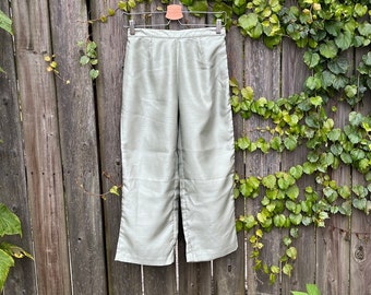 May include: Light green, wide-leg pants hanging on a wooden fence. The pants have a high waist and a subtle sheen. The background features a wooden fence covered in green vines and autumn foliage.
