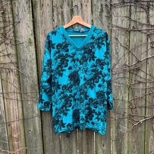 May include: A teal blue long-sleeved shirt with a black floral print. The shirt has a V-neck and is hanging on a wooden hanger.