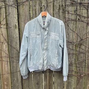May include: A light blue jacket with a zipper closure and a ribbed collar and cuffs. The jacket is hanging on a wooden hanger against a wooden fence.