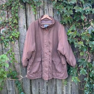 Puede incluir: Una chaqueta marrón con capucha, cremallera, cintura con cordón y dos bolsillos. La chaqueta está colgada de una percha de madera contra una valla de madera con enredaderas verdes.