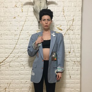 Könnte beinhalten: Eine Person trägt einen hellblauen Blazer mit bunten Patches, schwarze Leggings und blaue und gelbe Socken. Die Person steht vor einer weißen Wand mit einem weißen Tierschädel, der daran hängt.