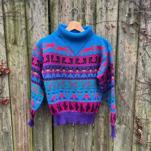 May include: A vintage turtleneck sweater with a vibrant pattern. The sweater features a blue turtleneck and cuffs, with a design in shades of purple, teal, and red. The pattern includes geometric shapes and stylized figures. The sweater is hanging on a wooden hanger.