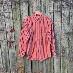 May include: A coral-colored button-down shirt with vertical black stripes. The long-sleeved shirt is hanging on a wooden hanger. The shirt has a classic collar and buttoned cuffs. The background is a weathered wooden fence.