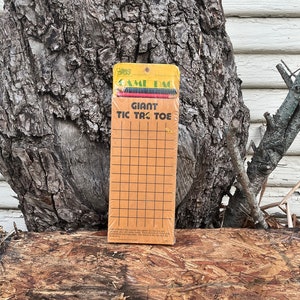 May include: A yellow and orange cardboard game box with the text "Giant Tic Tac Toe" on the front. The box is sitting on a piece of wood.