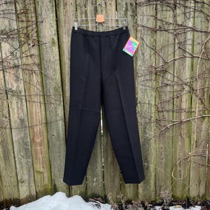 Puede incluir: Pantalón negro de pierna recta y cintura alta. El pantalón está colgado de una percha de madera contra una valla de madera.
