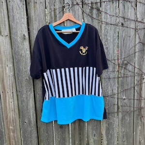 May include: Black and blue V-neck t-shirt with a nautical design. The shirt features a black upper section with a blue trim around the neckline, a white and black striped middle section, and a blue bottom section. A small emblem is on the upper left.