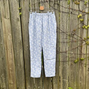 May include: Light blue pants with a white floral pattern hanging on a wooden hanger against a wooden fence.