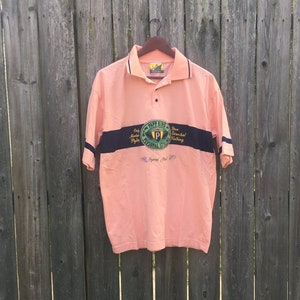 May include: A peach-colored polo shirt with a navy blue and white stripe across the chest. The shirt features a circular logo with the text "P.S. Personal Style" and a gold "P" inside a green circle. The shirt has a button-up collar and short sleeves.