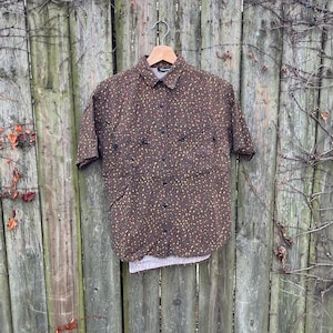 May include: Brown short-sleeve button-down shirt with a speckled pattern in yellow and brown. The shirt has a collar and two chest pockets. It is hanging on a wooden hanger against a weathered wooden fence.