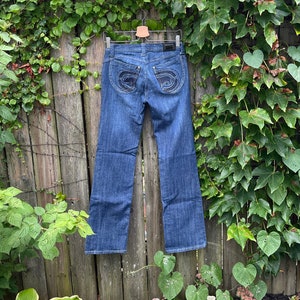May include: A pair of blue denim jeans hanging on a wooden fence. The jeans have a dark wash and a bootcut style. The back pockets have a unique design with a stitched circle.
